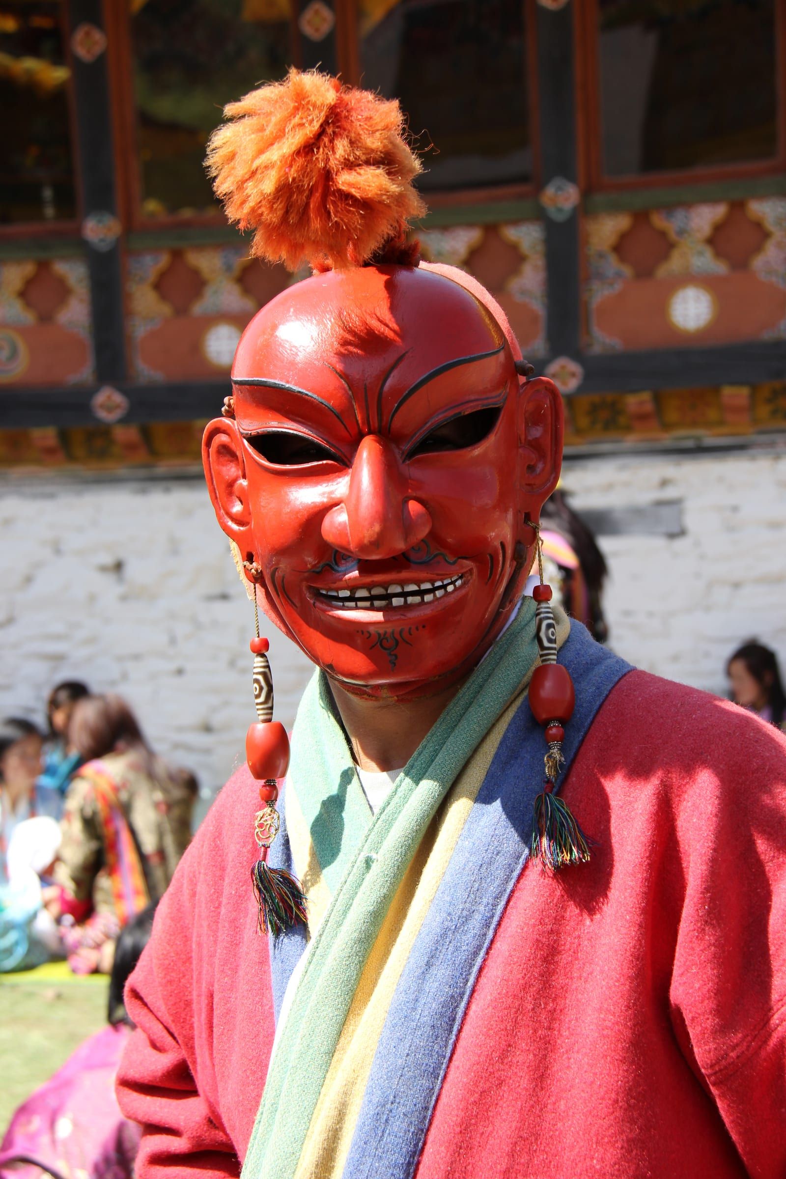An atsara in a red mask with a long nose, a tuft of orange hair and beaded earrings.