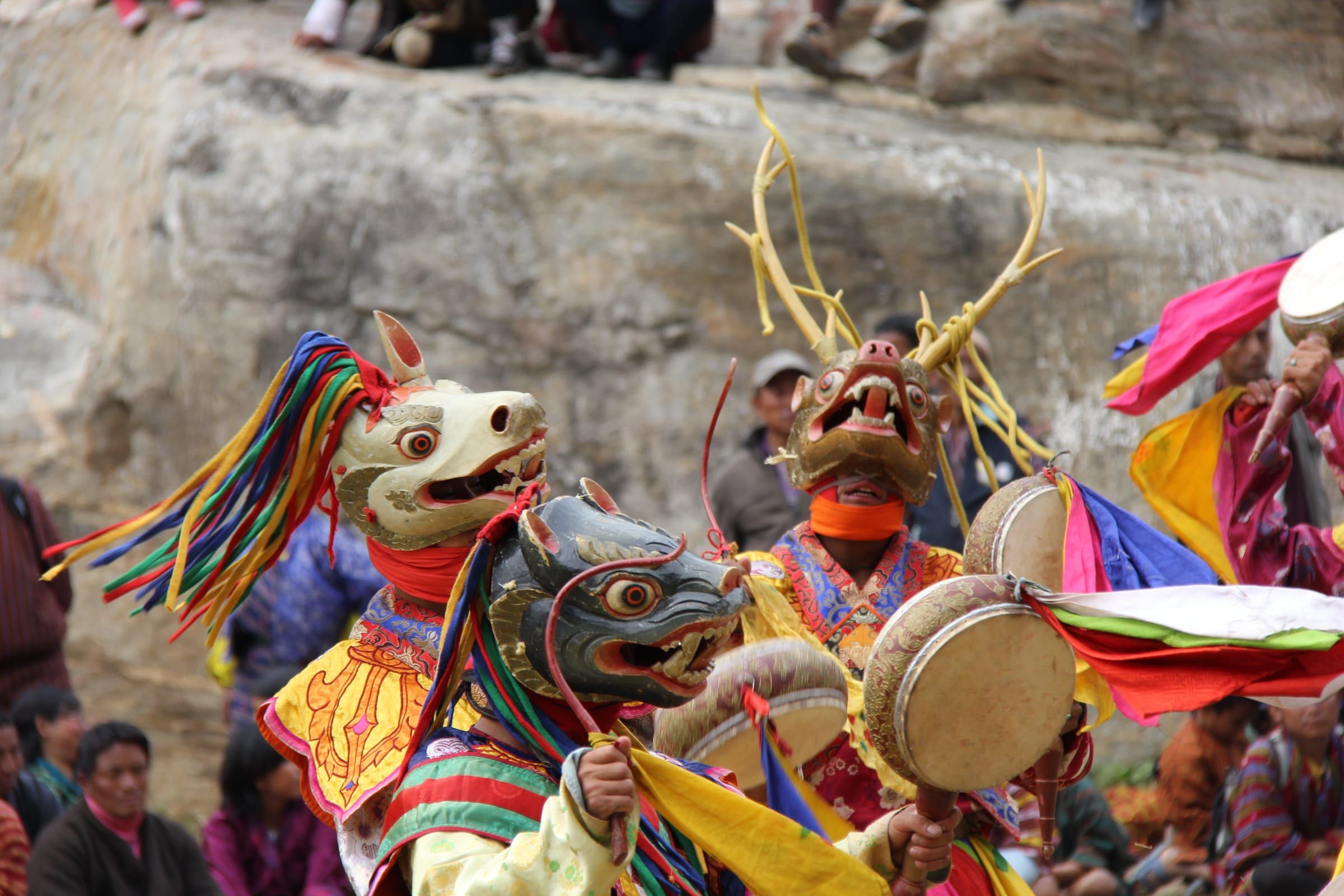Dancers in animal masks, one with antlers, swing hand drums in front of a rock face.