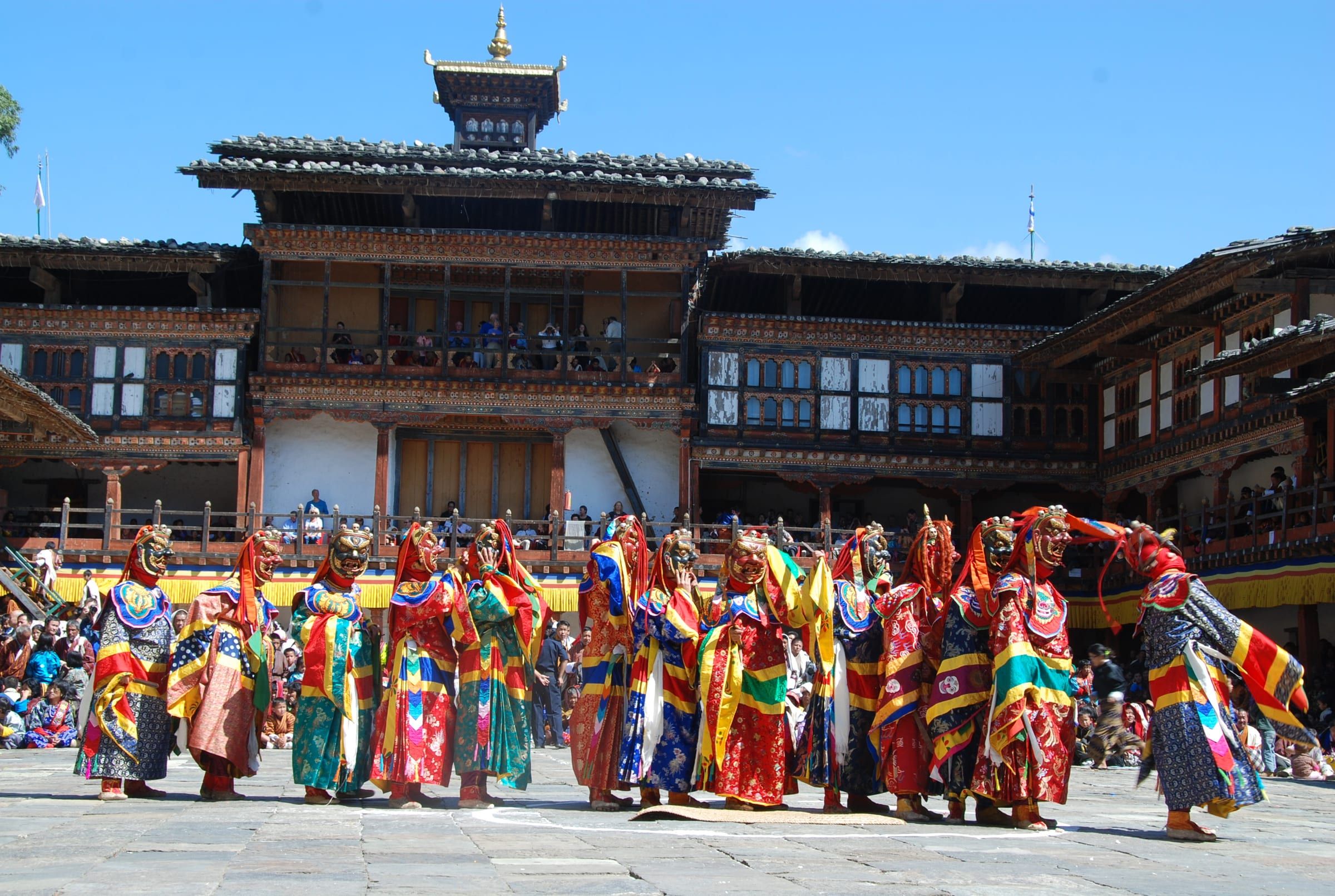 A line of masked dancers in brocade robes in a dzong courtyard, below wooden galleries.