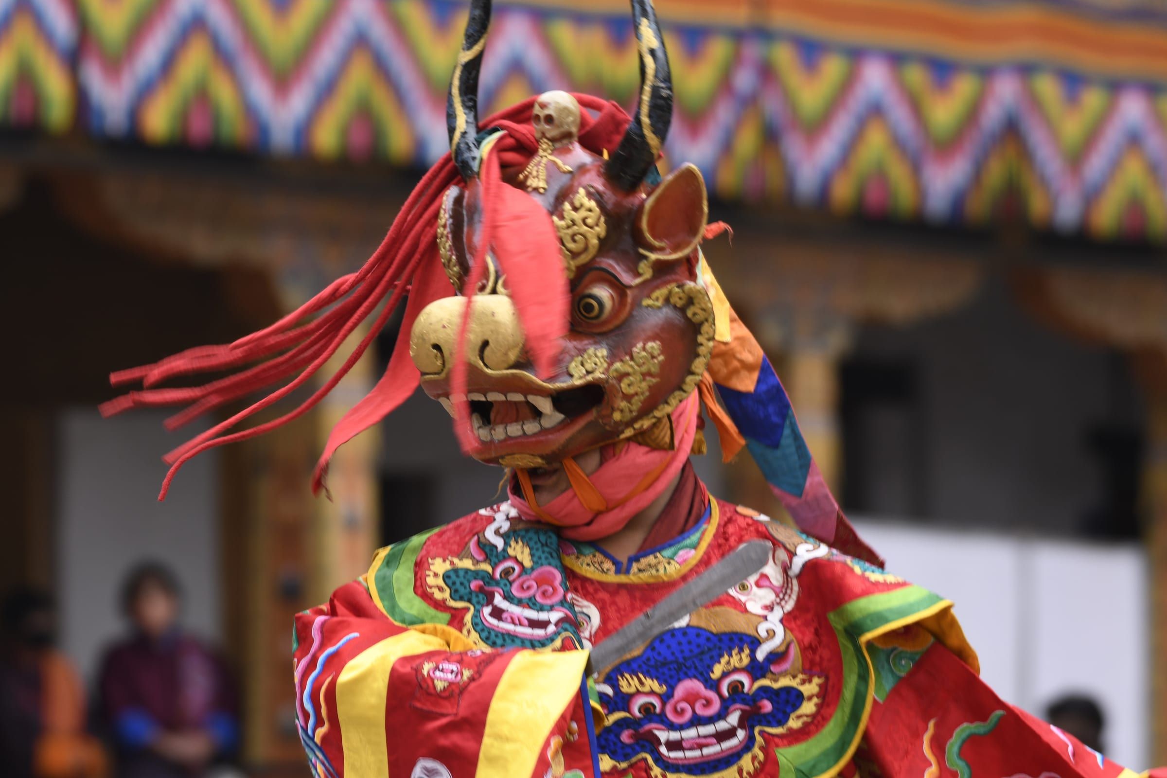 A dancer in a horned bull mask topped with a small skull turns in red brocade robes.